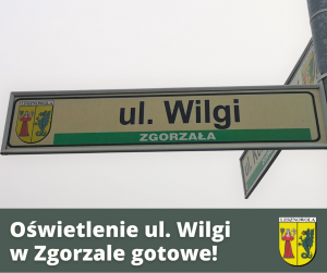 Biały napis "Oświetlenie ul. Wilgi w Zgorzale gotowe!" i Herb Gminy Lesznowola na ciemny tle. Znak z nazwą ulicy.