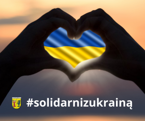 Biały napis "#solidarnizukrainą". W centralnej części zdjęcia dłonie ułożone w kształcie serca na fladze Ukrainy