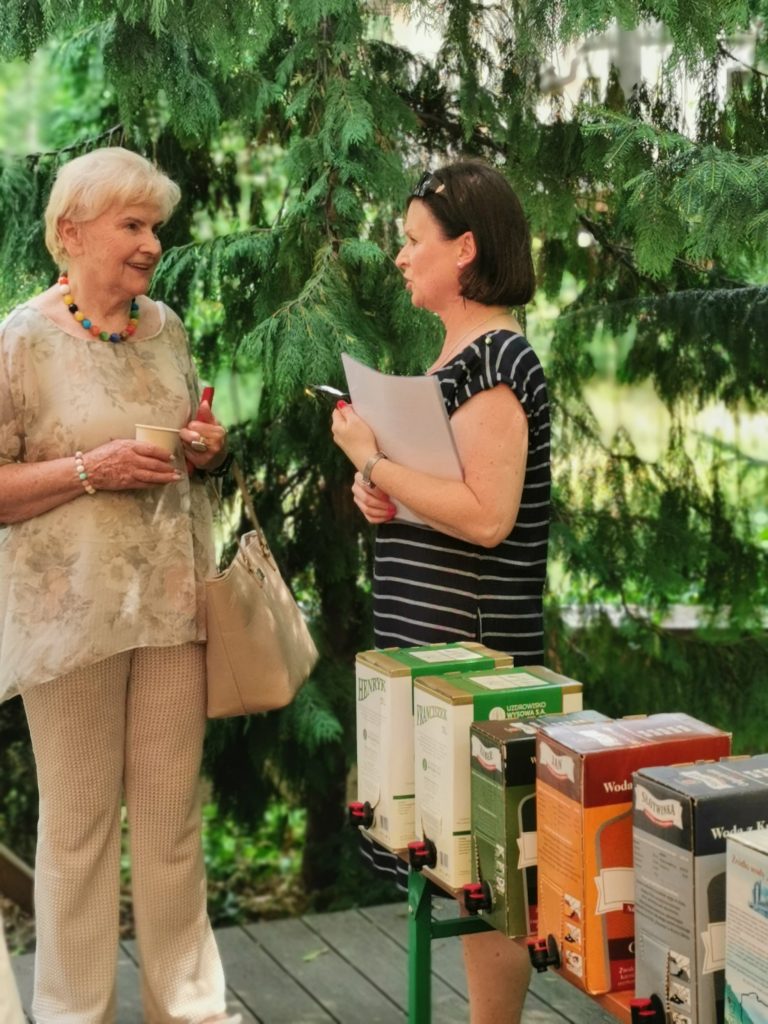 Stoją dwie kobiety, rozmawiają. Po prawej widać kolorowe pudełka.