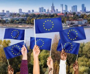 Sześć rąk podnosi flagi UE. W tle widać Warszawę.