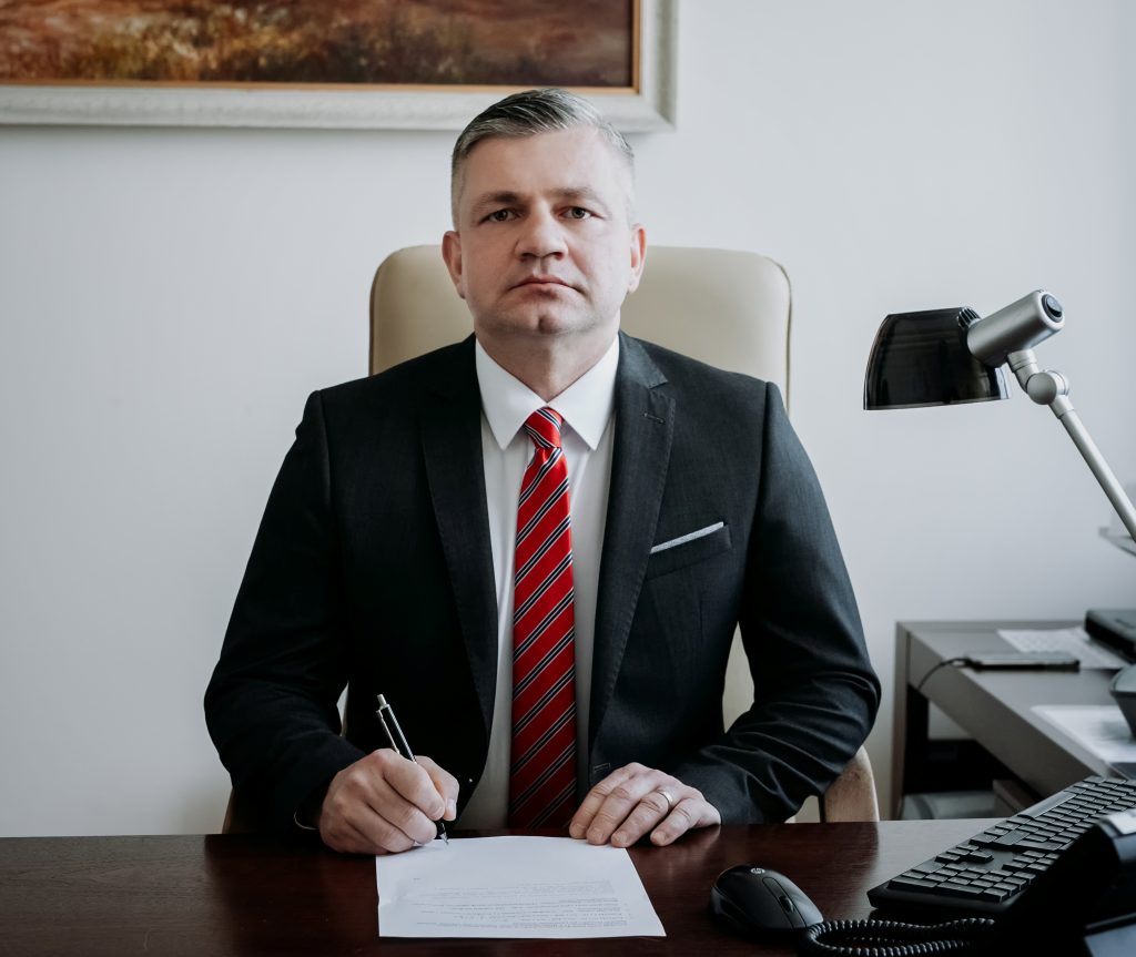 mężczyzna w ciemnym garniturze siedzi za stołem i opiera dłonie o stół, na stole kartak i d