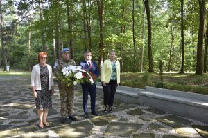 Las, cztery osoby stoja , osoba w mundurze trzyma wienieć z kwiatami.