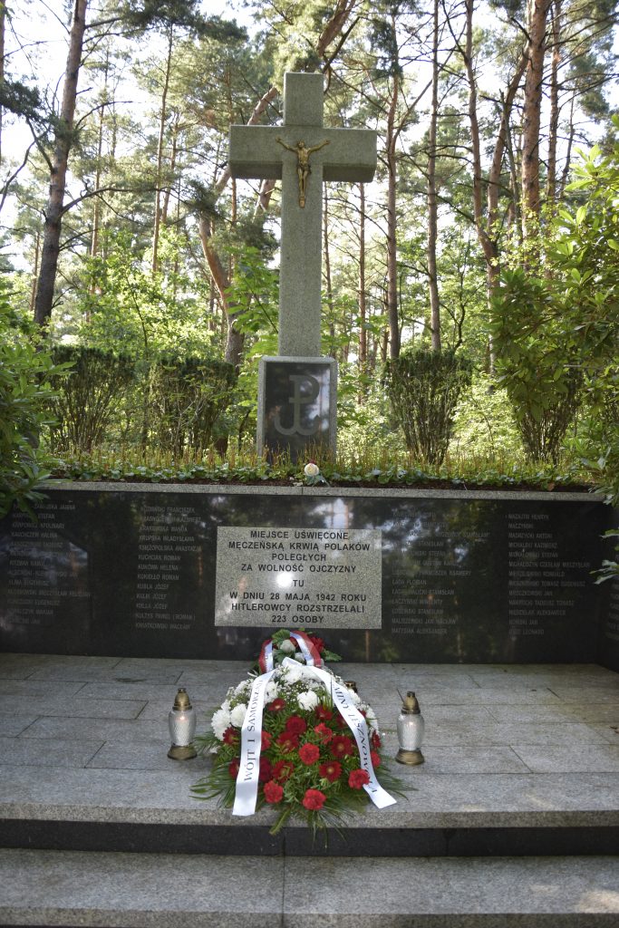 Wysoki kamienny krzyż w lesie. Na pierwszym planie biało-czerwona wiązanka kwiatów i dwa znicze.