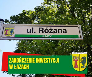 Drzewo, tablica z nazwą ulicy: ul. Różana. Poniżej na zielonym pasku napis: ZAKOŃCZENIE INWESTYCJI W ŁAZACH. N a dole po prawej herb Gminy.