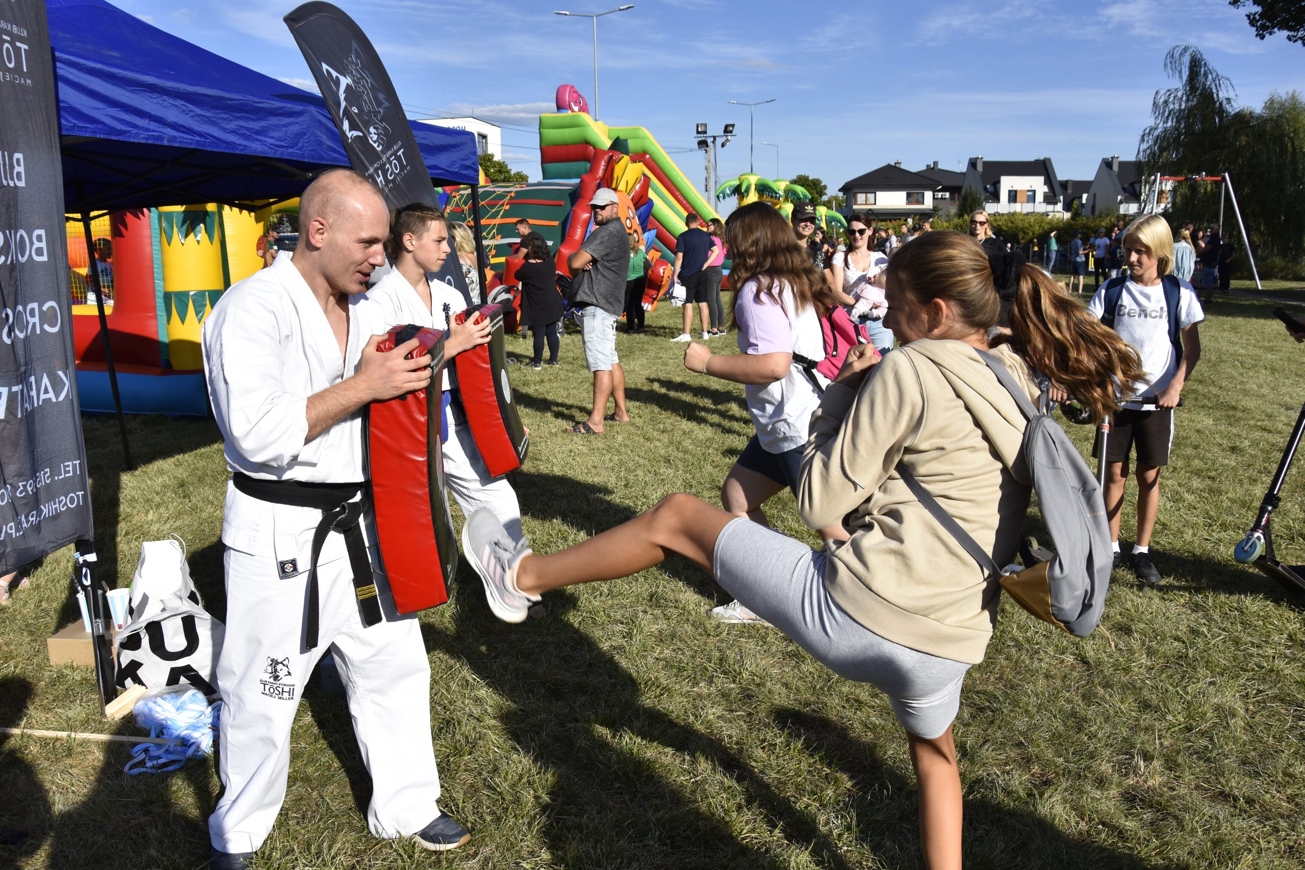 Teren zielony, ludzie. Na pierwszym planie po lewej stronie mężczyzna w białym kostuimie trzyma tarcze, w ktorą kopie dziewczyna po prawej.