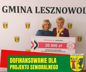 Dwie kobiety stoja na tle białej ścianki z herbami i napisem Gmina Lesznowola, trzymaja w ręce tablice informacyjną.