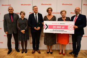 Na tle ściany stoi 5 osób,Pierwsza, druga i trzecia od lewej trzymają prostokątna tablice.