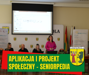 U góry nad sufitem zawieszony jasny ekran, za stołem ludzie, jedna osoba stoi. na zielonym pasku napis: APLIKACJA I PROJEKT SPOŁECZNY - SENIORPEDIA, obok herb gminy