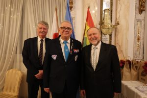 Trzech mężczyzn pozuje do zdjęcia. W tle znajdują się flagi Polski, ONZ i Lesznowoli.