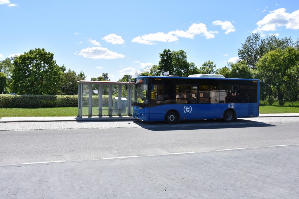 Niebieski autobus podjeżdża na przystanek. Pogoda jest słoneczna.