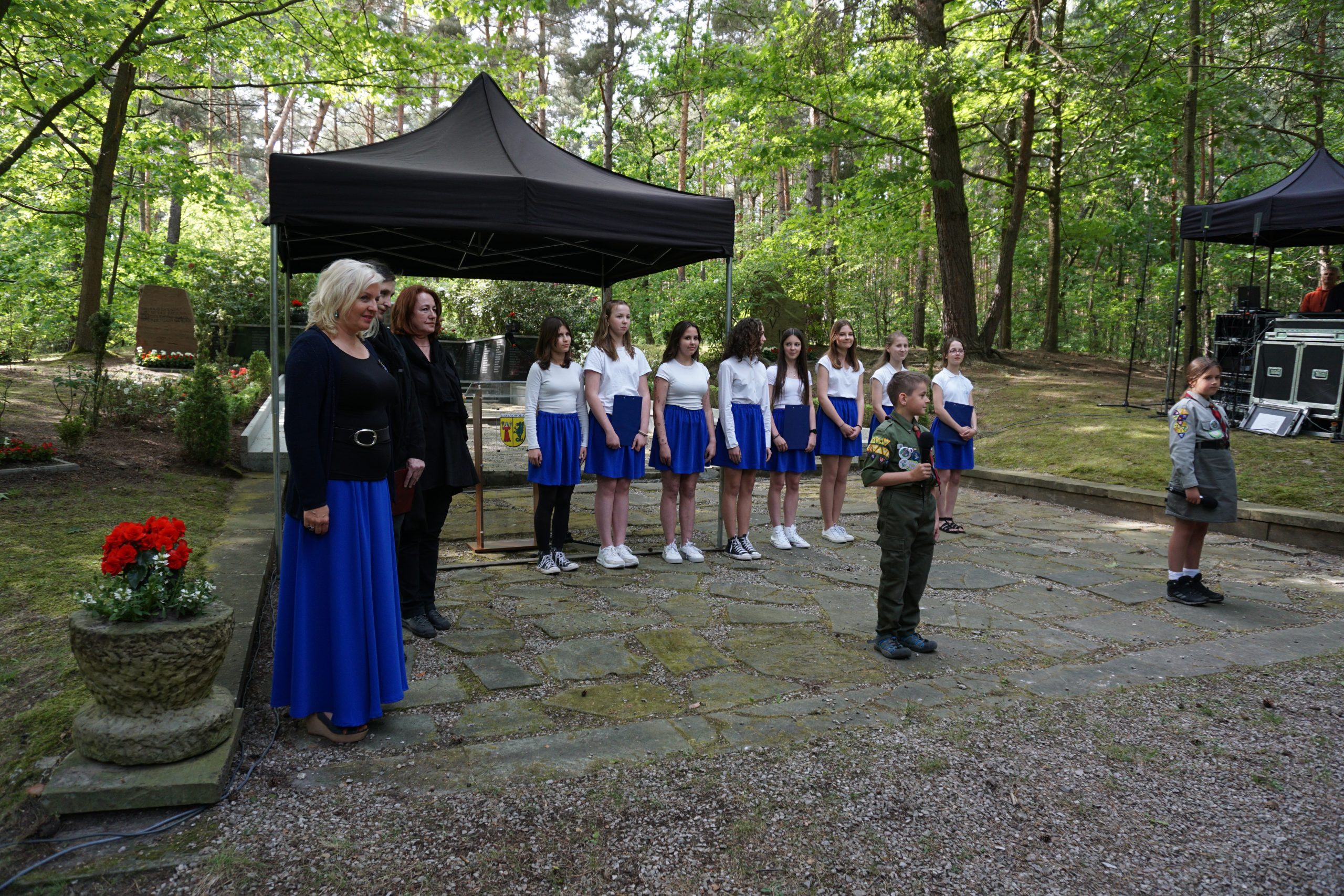 chór, dziewczynki ubrane w granatowe spódnice i białe bluzki, dwie kobiety oraz chłopczyk