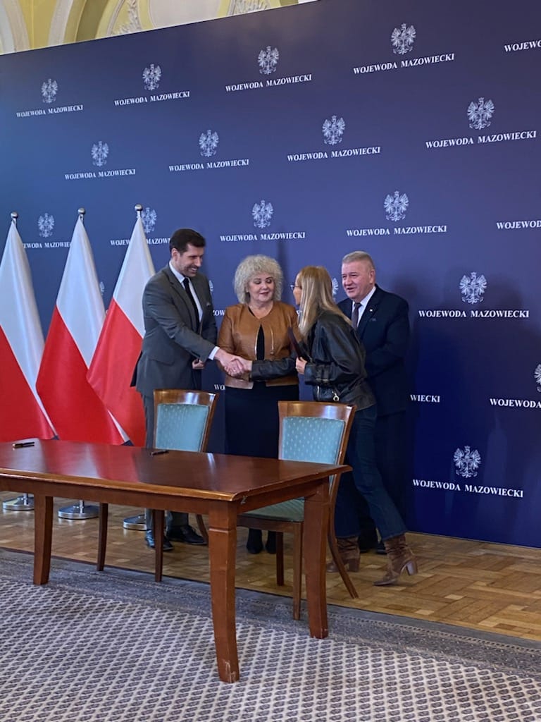 4 osoby w Urzędzie Marszałkowskim podczas podpisywania umowy, na tle niebieskiej ścianki promocyjnej i flag państwowych.