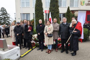 Grupa mężczyzn i kobiet stoi pod pomnikiem, za nimi urząd