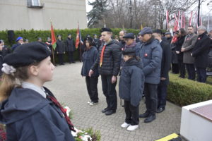Grupa mężczyzn z harcerzami składa kwiaty pod pomnikiem. Na drugim planie widoczne inne osoby.