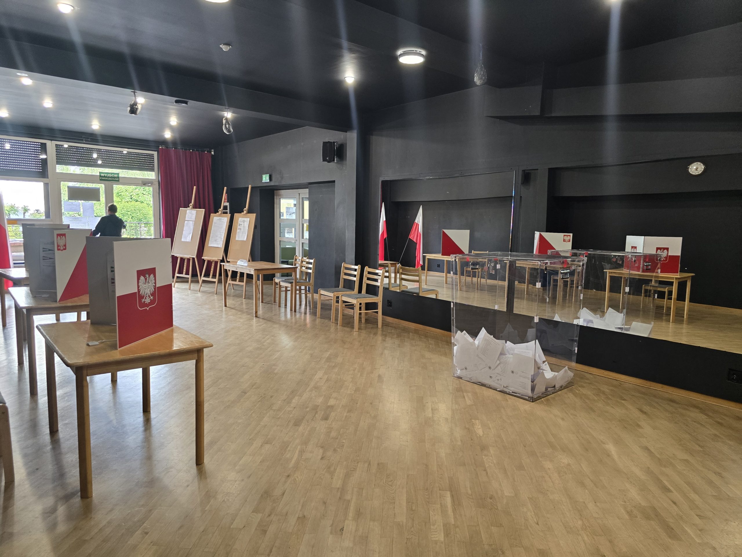 Na zdjęciu lokal wyborczy, pomieszczenie o czarnych ścianach z duzym lustrem. Widac stanowiska do głosowania i urnę wyborcza obok lustra.