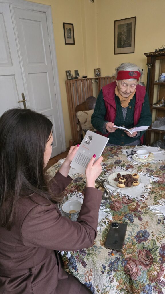 Na zdjęciu dwie kobiety, jedna widoczna z tyłu - trzyma w ręku książkę