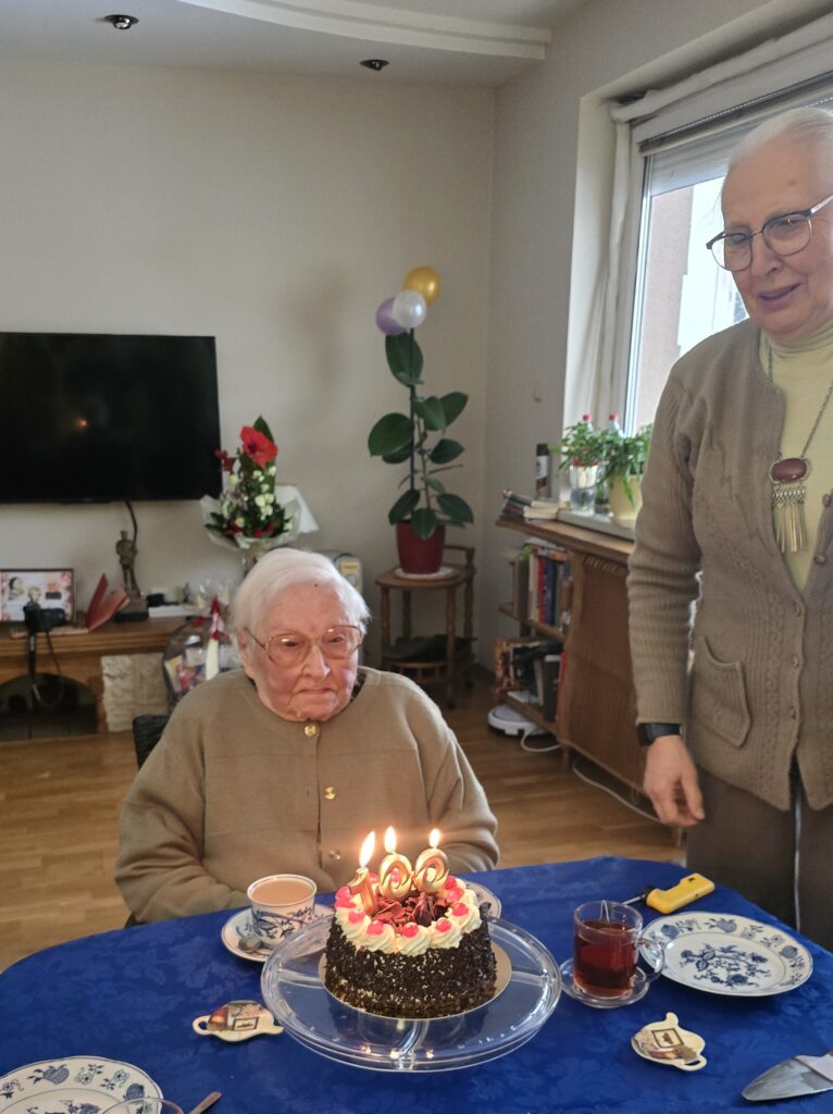Starsza pani siedzi przy stole nakrytym niebieskim obrusem, przed nią tort z zapalonymi świeczkami. Obok stoi kobieta.