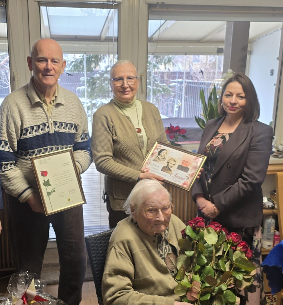 Na zdjęciu trzy osoby stojące na tle okana, pozują do zdjęcia, nieco przed nimi starsza pani z siedząca na krześle z bukietem róż w rękach.